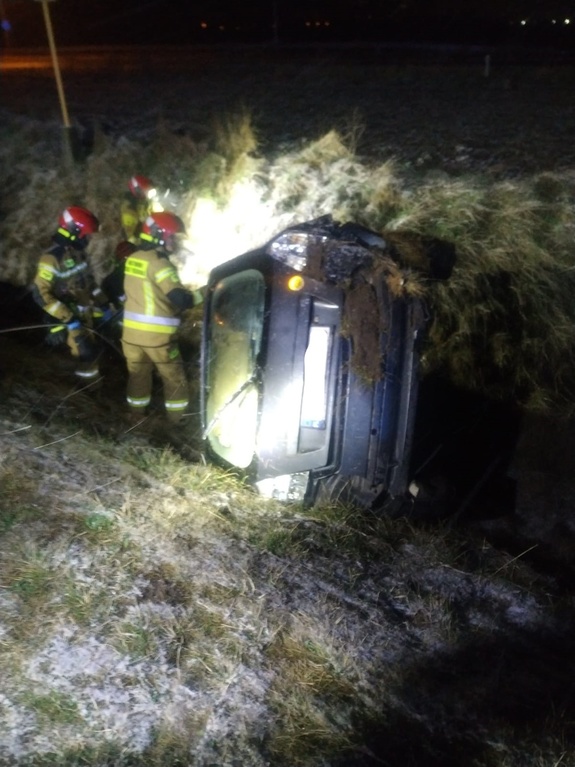 Zdjęcie kolorowe wykonane w porze nocnej-przedstawia samochód m-ki fiat stilo w kolorze granatowym. Samochód jest przewrócony na bok , obok pojazdu znajdują się strażacy