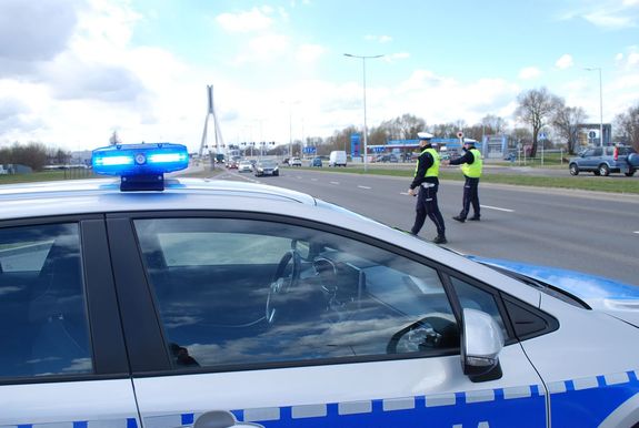 na zdjęciu umundurowani policjanci zatrzymują kierującego do kontroli. Na pierwszym planie widać radiowóz, za nim na jezdni stoją policjanci drogówki podający tarczą do zatrzymywania sygnał kierującemu. W tle widać samochody poruszające się ul. Rzecha i most Tadeusza Mazowieckiego.