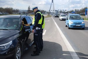 Policjanci drogówki stoją przy zatrzymanym samochodzie i sprawdzają trzeźwość kierującego. W tel widać samochody poruszające się ul. Rzecha i most Tadeusza Mazowieckiego.