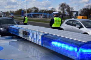 Na pierwszym planie policyjna belka radiowozu z włączonymi sygnałami świetlnymi. W tle policjanci zatrzymujący pojazdy.