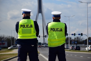 W środku kadru dwaj policjanci ruchu drogowego (widoczni od tyłu). W tle most.