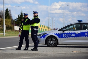 Dwaj policjanci ruchu drogowego w kamizelkach (po lewej) stoją przy drodze, obok, po prawej radiowóz.