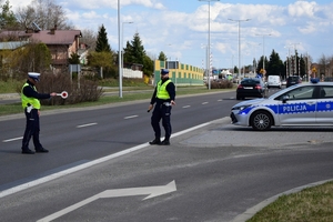 Dwaj policjanci ruchu drogowego w kamizelkach (po lewej) przy drodze, zatrzymują pojazd. Obok, po prawej radiowóz.