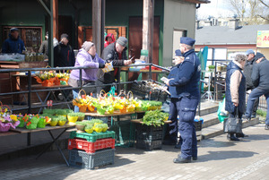 Zdjęcie kolorowe wykonane w porze dziennej przedstawia bazar warzywny przy ul. Sportowej w Przemyślu. W tle widoczni są dzielnicowi umundurowani , którzy podczas służby informują kupujących jak i sprzedających o zachowaniu ostrożności podczas zakupów przedświątecznych.