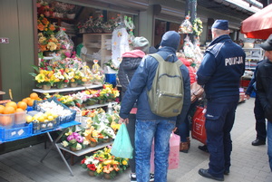 Zdjęcie kolorowe wykonane w porze dziennej przedstawia bazar warzywny przy ul. Sportowej w Przemyślu. W tle widoczni są dzielnicowi umundurowani , którzy podczas służby informują kupujących jak i sprzedających o zachowaniu ostrożności podczas zakupów przedświątecznych.