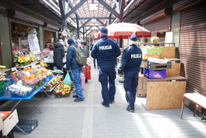 Zdjęcie kolorowe wykonane w porze dziennej przedstawia bazar warzywny przy ul. Sportowej w Przemyślu. W tle widoczni są dzielnicowi umundurowani , którzy podczas służby informują kupujących jak i sprzedających o zachowaniu ostrożności podczas zakupów przedświątecznych.