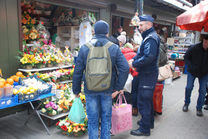 Zdjęcie kolorowe wykonane w porze dziennej przedstawia bazar warzywny przy ul. Sportowej w Przemyślu. W tle widoczni są dzielnicowi umundurowani , którzy podczas służby informują kupujących jak i sprzedających o zachowaniu ostrożności podczas zakupów przedświątecznych.