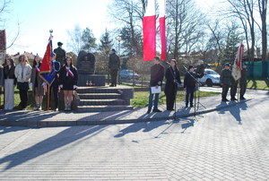 Na zdjęciu przed obeliskiem upamiętniającym ofiary zbrodni katyńskiej stoją dwa poczty sztandarowe, po lewej stronie poczet szkolny po prawej poczet strzelca. Przed obeliskiem dwoje strzelców wystawi wartę. Przed pomnikiem widać młodzież uczestnicząca w obchodach