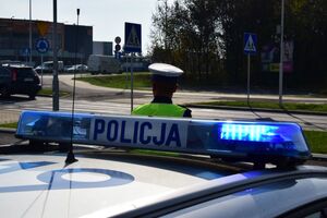 Zdjęcie kolorowe, dzień słoneczny, na pierwszym planie belka radiowozu, przed pojazdem stoi policjant ruchu drogowego - w białej czapce i odblaskowej kamizelce. W tle jadące samochody