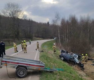 Na zdjęciu widać służby pracujące na miejscu wypadku drogowego. W centralnej części zdjęcia widać przyczepkę stojącą na jezdni