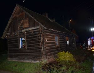 Na zdjęciu w nocy na pierwszym planie widać drewniany budynek, w którym doszło do pożaru, w tle za budynkiem stoją częściowo widoczne oświetlone wozy strażackie.