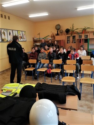 Policjanci podczas spotkania z dziećmi na dniach otwartych. W środku kadru stoi twarzą do uczniów, którzy siedzą na krzesłach. Na pierwszym planie policyjny sprzęt - kask i elementy umundurowania.