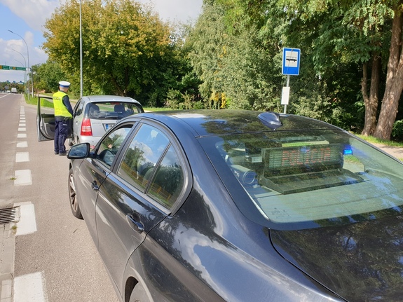 policjanci podczas kontroli drogowej, na fotografii nieoznakowany pojazd służbowy policji, tuż przed nim osobowy samochód zatrzymany do kontroli oraz umundurowany policjant wydziału ruchu drogowego