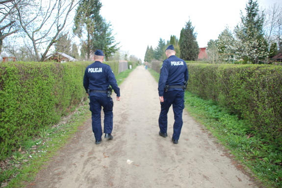 Zdjęcie kolorowe wykonane w porze dziennej -przedstawia ogródki działkowe na terenie miasta Przemyśl. Dwóch policjantów patroluje ogródki oraz rozmawia z działkowcami o bezpieczeństwie