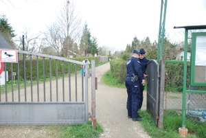 Zdjęcie kolorowe wykonane w porze dziennej -przedstawia ogródki działkowe na terenie miasta Przemyśl. Dwóch policjantów patroluje ogródki oraz rozmawia z działkowcami o bezpieczeństwie