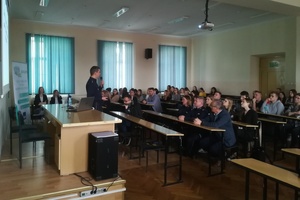 Policjant prowadzi spotkanie ze studentami. Policjant stoi w centrum kadru przy biurku,  twarzą do studentów.