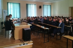 Po lewej przy biurku siedzą studenci, po prawej w tle sala, w ławkach siedzą studenci oraz troje policjantów.