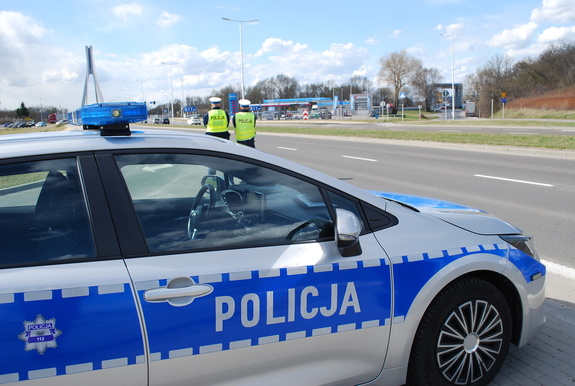 Na zdjęciu widać radiowóz z napisem policja i dwóch umundurowanych funkcjonariuszy w kamizelkach odblaskowych, w tle ulica i stacja paliw.