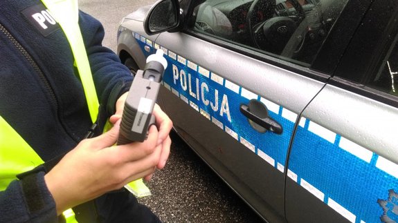Policjant trzyma w rękach alkomat. W tle radiowóz.