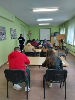 Na zdjęciu znajduje się młodzież siedząca w ławkach w sali lekcyjnej oraz umundurowany policjant