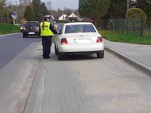 Na zdjęciu policjantka ruchu drogowego, która dokonuje badania trzeźwości kierującego białym audi. W tle droga i jadące po niej pojazdy.
