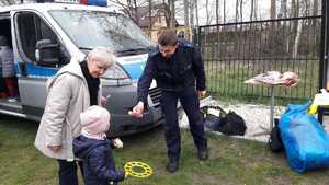 na zdjęciu policjant na tle radiowozu dający element odblaskowy dziecku