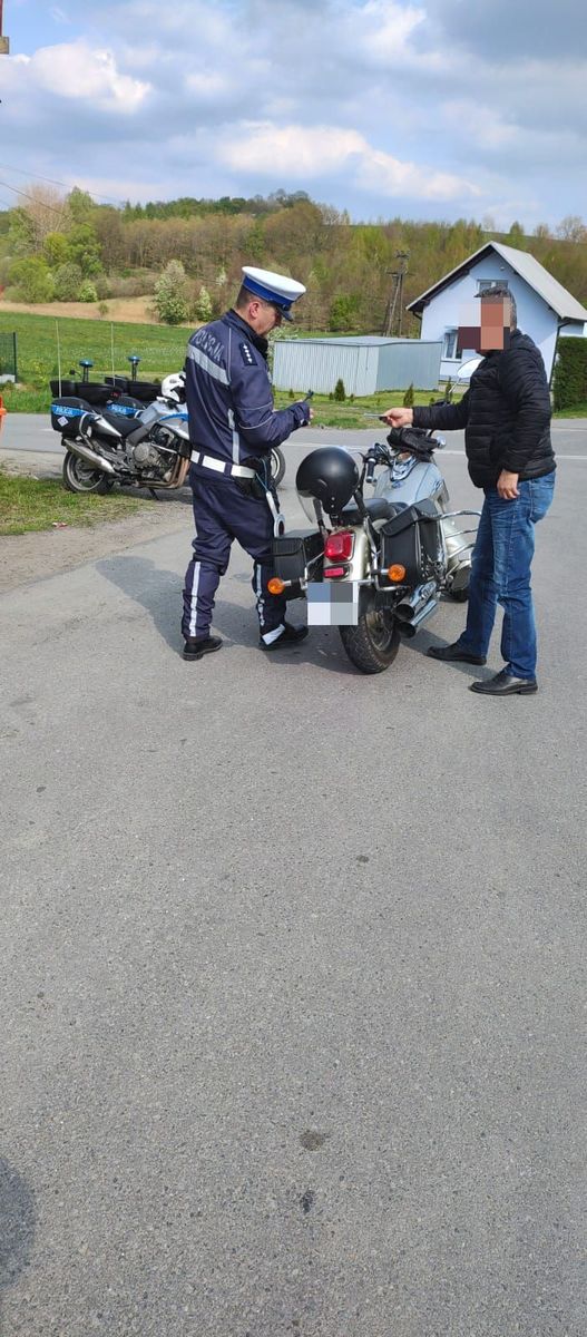Policjant ruchu drogowego, kontroluje kierującego motocyklem. Obaj stoją przy pojeździe osoby kontrolowanej.