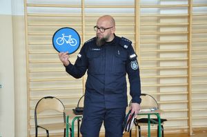 Na zdjęciu umundurowany policjant podczas spotkania w sali gimnastycznej uczy dzieci o znakach drogowych. Stojący przodem Policjant pokazuje uczniom znak droga dla rowerów.