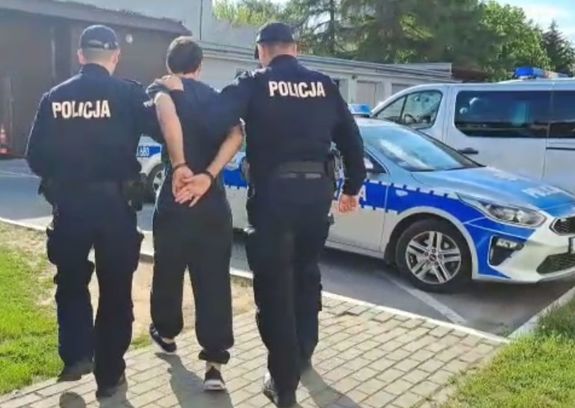 Dwóch policjantów prowadzi zatrzymanego mężczyznę, na drugim planie radiowóz