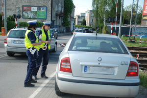 Zdjęcie kolorowe wykonane w porze dziennej  przedstawia przejazd kolejowy z zaporami na którym patrol policji dokonuje kontroli kierowcy