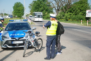 Zdjęcie kolorowe wykonane w porze dziennej  przedstawia przejazd kolejowy bez zapór na którym patrol policji dokonuje kontroli rowerzysty