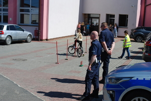 Na pierwszym planie policjanci ruchu drogowego podczas gminnych kwalifikacji turnieju. W oddali uczeń szkół, jadąc rowerem pokonuje tor przeszkód.