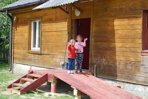 Zdjęcie kolorowe wykonane w porze dziennej –przedstawia dom w którym na jego podjedzie stoją dwie małe dziewczynki. Dzieci machają rączkami na pożegnanie policjantów