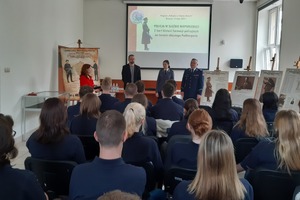 Policjanci podczas warsztatów. Na pierwszym planie uczniowie (widok od tyłu) siedzący na krzesłach. W centrum kadru policjanci stojący twarzą do uczniów. Za nimi ekran z prezentacją.