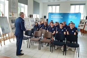 Policjanci podczas warsztatów. Na pierwszym planie uczniowie (widok od tyłu) siedzący na krzesłach. W centrum kadru policjanci stojący twarzą do uczniów. Za nimi ekran z prezentacją.
