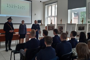 Policjanci podczas warsztatów. Na pierwszym planie uczniowie (widok od tyłu) siedzący na krzesłach. W centrum kadru policjanci stojący twarzą do uczniów. Za nimi ekran z prezentacją.