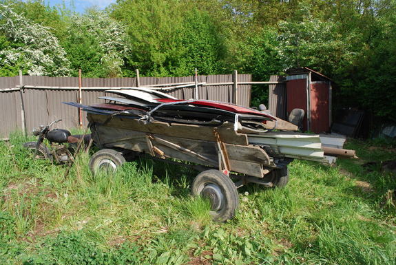 Zdjęcie kolorowe wykonane w porze dziennej przedstawia prywatna posesję na której widoczny jest wóz konny wypełniany elementami metalowymi. Wóz podpięty jest pod stary motor w kolorze czarnym