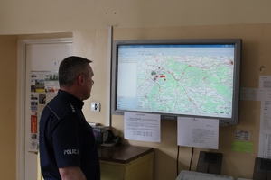 Dyżurny jednostki Policji patrzy na wiszący przed nim na ścianie ekran, na którym wyświetla się mapa.