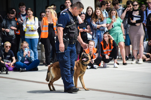 Na pierwszym planie policjant z psem służbowym na placu przed budynkiem. W tle uczniowie i studenci.