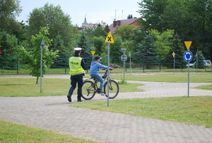 Na zdjęciu umundurowana funkcjonariusza drogowego w żółtej odblaskowej kamizelce ocenia przejazd jednego z uczestników. Obok niej jedzie na rowerze jeden z uczniów sprawdzianu.
