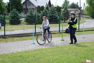 Na zdjęciu umundurowana funkcjonariusza drogowego w żółtej odblaskowej kamizelce ocenia przejazd przez tor przeszkód. Obok niej jedzie na rowerze dziewczynka reprezentująca jedną ze szkół.