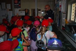 na zdjęciu policjant opowiada dzieciom o pracy dyżurnego komendy. W tel widać stanowisko kierowania rzeszowskiej komendy. Przed nim stoi grupa dzieci w czerwonych czapeczkach i słucha opowiadania policjanta.