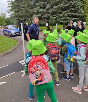 policjanci podczas prelekcji dot. bezpieczeństwa wśród dzieci w zielonych kapeluszach. W tle policyjny radiowóz