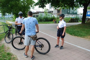 Na zdjęciu, na alejce na Bulwarach policjantka i policjant z patrolu rowerowego legitymują rowerzystę. Policjanci ubrani są w białe koszulki polo z krótkim rękawem z napisem POLICJA na lewej piersi i prawym rękawie koszulki, czarne krótkie spodnie i czarne buty. Na głowie mają czarne kaski. Legitymowany mężczyzna stoicy tyłem, obok niego rower ubrany w niebieską koszulkę z krótkim rękawem i krótkie spodnie koloru szarego. W tle drzewa i plac budowy.