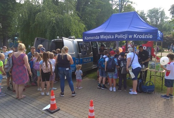 Niebieski namiot profilaktyczny z napisem Komenda Miejska Policji w Krośnie, wokół dzieci i stojący obok radiowóz