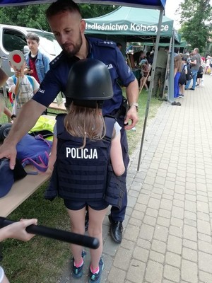 Policjant prezentujący wyposażenie służbowe, stojąca przed nim dziewczynka w elementach policyjnego umundurowania