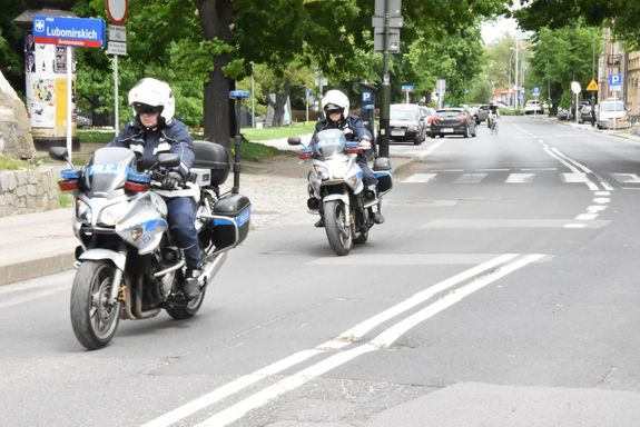 Na zdjęciu jadące ulicą Szopena dwa motory policyjne, na których  siedzą policjanci ubrani w białe kaski na głowach i czarne kombinezony. W tle drzewa, zaparkowane samochody i budynku.