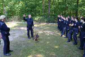Na zdjęciu znajduje się dwoje policjantów, pies służbowy oraz uczniowie klas o profilu policyjnym stojący w kompleksie leśnym