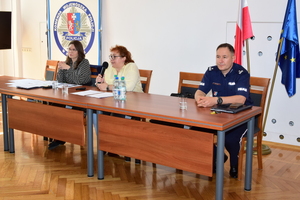 W centrum kadru, przy stole siedzą trzy osoby. Od prawej - umundurowany policjant, w środku kobieta w jasnej bluzce, trzyma w rękach mikrofon, po lewej kobieta w czarno-białej bluzce. W tle, po prawej dwie flagi - biało-czerwona i flaga UE. W tle po lewej logo z policyjną gwiazdą, herbem Podkarpacia i napisami Komenda Wojewódzka Policji w Rzeszowie oraz Policja.