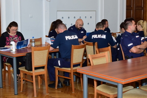 Uczestnicy szkolenia w trakcie spotkania. Po lewej kobieta w okularach trzyma w rękach broszurę informacyjną, w środku i po prawej umundurowani policjanci i policjantki (tyłem i przodem), siedzący przy stołach i czytający broszury informacyjne.